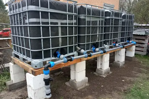Kilka połączonych zbiorników IBC na drewnianej konstrukcji z systemem rur i zaworów.