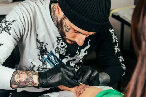 Artysta z tatuażami na twarzy i rękach tworzy nowy wzór na przedramieniu klienta. Dyskutują o cukrzycy a tatuażu.