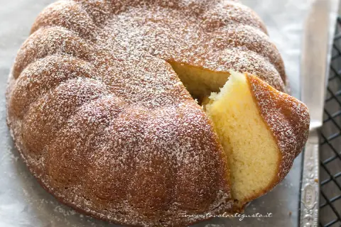 Una soffice torta allo yogurt, cosparsa di zucchero a velo, con una fetta tagliata che ne rivela l'interno dorato e profumato.