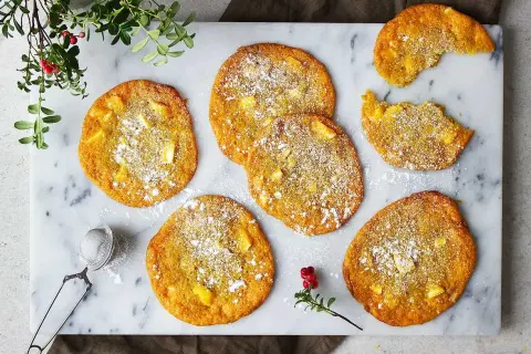 Sju gyllene saffranscookies med vit choklad, pudrade med florsocker, ligger på ett marmorfat. En bit är tagen från en cookie.