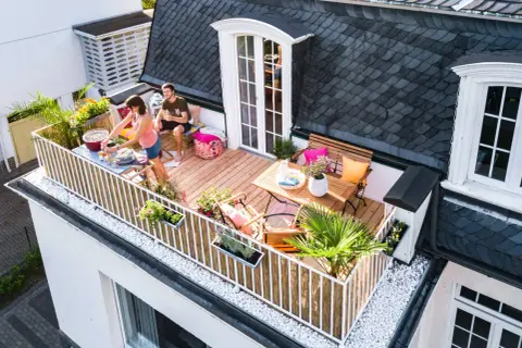 Ein Paar genießt ein Grillfest auf ihrem neu gestalteten Balkon. Der Balkon ist mit Holzmöbeln, Pflanzen und Dekorationen geschmückt.