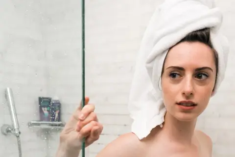 Ragazza con asciugamano in testa guarda preoccupata la doccia, pensando che la paraffina liquida fa male alla pelle.