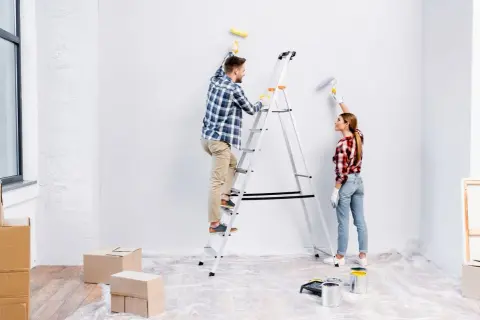 Para odnawia mieszkanie. Mężczyzna na drabinie maluje ścianę, kobieta obok też maluje.