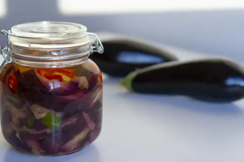 Berenjenas en escabeche, cortadas y marinadas en un frasco de vidrio, con berenjenas frescas desenfocadas al fondo.