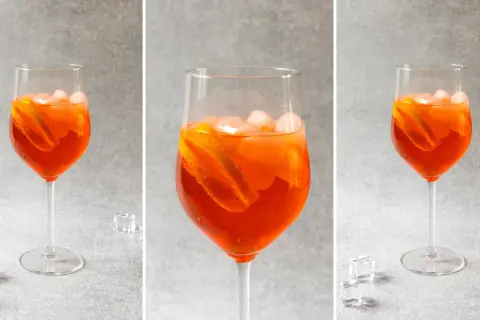 Trzy identyczne kieliszki z pomarańczowym drinkiem Aperol Spritz, lodem i plasterkami pomarańczy na szarym tle.