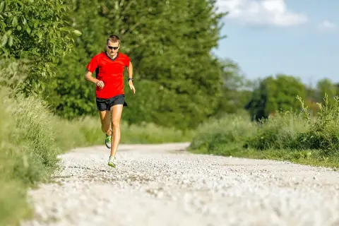 Mann in rotem Shirt und schwarzen Shorts absolviert einen Fartlek Lauf auf einem Schotterweg, umgeben von grüner Natur.