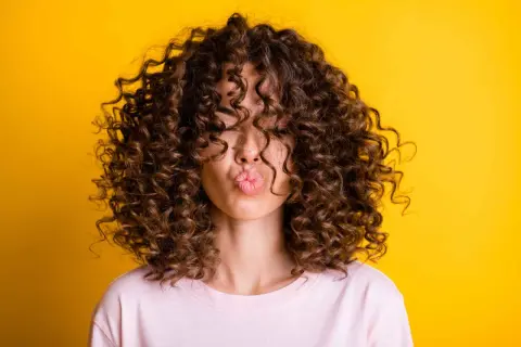 Mujer con un espectacular pelo rizado fino y castaño, haciendo un beso contra un fondo amarillo vibrante.