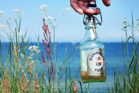 Eine Flensburger Gold Flasche wird vor der Küste gehalten. Der leichte Bier Geschmack passt perfekt zum Sommer.