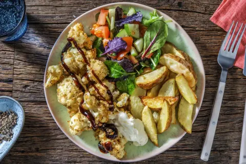 En tallrik med rostad blomkål i ugn, klyftpotatis och en fräsch sallad. Blomkålen är täckt med ströbröd och ringlad med en mörk sås.