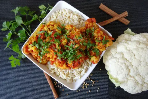 Aksamitne curry z kalafiora podane z ryżem, udekorowane świeżą natką pietruszki i czarnuszką. Obok leży świeży kalafior i laski cynamonu.
