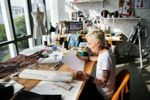 A designer sketches at a table filled with fabric and patterns, a scene that captures the essence of what is an artist residency: a dedicated space for creative exploration and development.