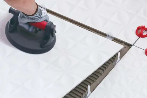 Mano con guante usa ventosa para colocar baldosas blancas. Herramientas alicatador en acción, con niveladores y adhesivo visible.