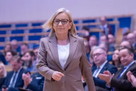 Kobieta w okularach i brązowej marynarce idzie na tle publiczności.
