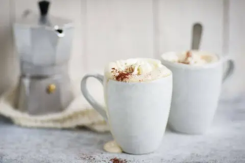 Två muggar med krämig mocca kaffe, toppad med vispgrädde och kakao. En mokabryggare står i bakgrunden, redo för ett nytt mocca kaffe recept.