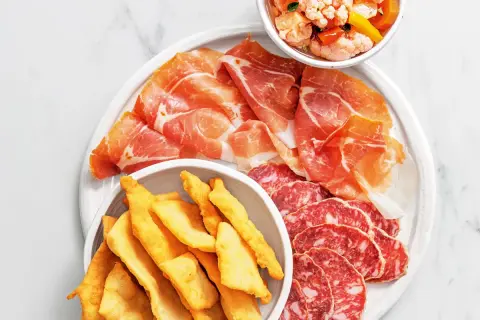 En tallrik med italienska delikatesser: tunt skivad prosciutto, salami och krispiga gnocco fritto. En skål med marinerade grönsaker.