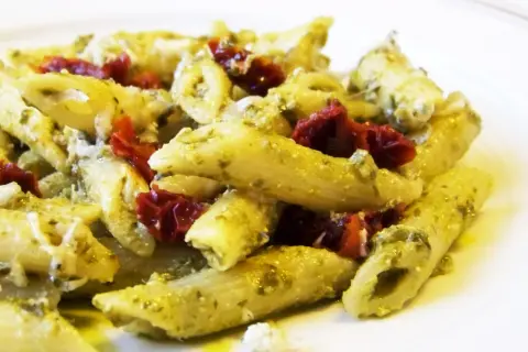 Makaron penne z zielonym pesto i suszonymi pomidorami na białym talerzu.