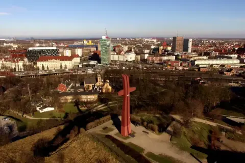 Wielki czerwony krzyż na górze gradowej góruje nad miastem. W tle widać zabudowania i drapacze chmur.