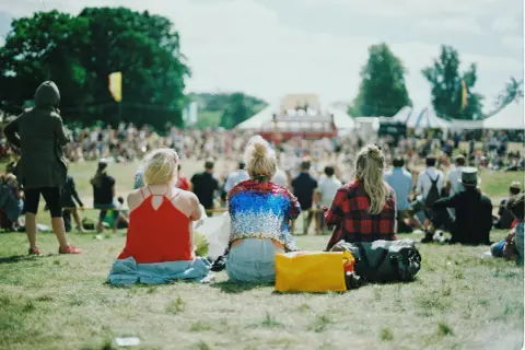 Festivalgäster sitter på gräset med en packlista i åtanke. En gul väska och en stor bag ligger redo för äventyr.