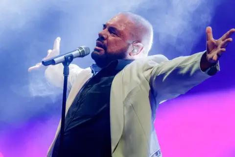 Wokalista wykonuje utwory Alphaville, w białej marynarce, z rękami uniesionymi w geście.