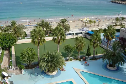 Vista aérea de un resort con piscina, cancha de césped, palmeras y una playa con el mar azul. Un lugar perfecto para sol y mar spa.