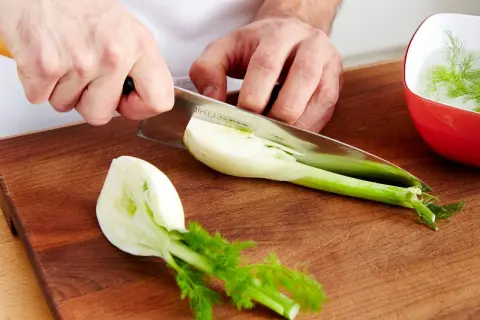 Hände schneiden Fenchel für leckere Fenchel Rezepte. Ein Teil liegt auf dem Holzbrett, ein anderer in einer roten Schüssel mit Wasser.