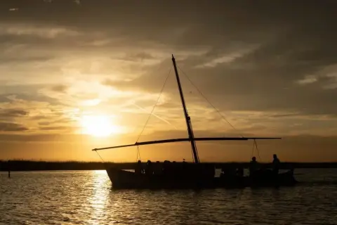 Un paseo en barca por la Albufera al atardecer, con el sol tiñendo el cielo de oro y las siluetas de los pasajeros reflejándose en el agua.