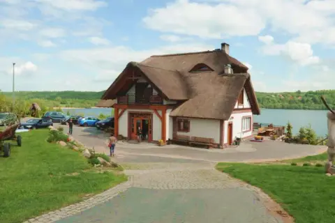 Dom z piękną, naturalną strzechą na dachu, położony nad jeziorem. Wokół zielony trawnik, drewniany wóz i rzeźby.