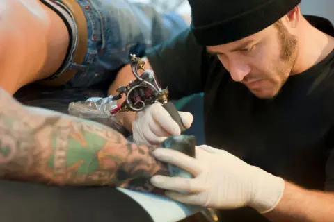 Artista tatuador concentrado, usando la mejor tinta para tatuajes en un brazo con diseños coloridos.