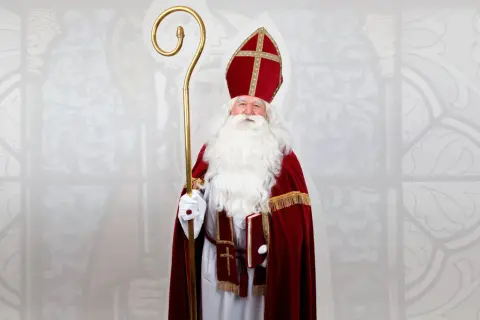 Mężczyzna w stroju św. Mikołaja biskupa, z białą brodą, mitrą i pastorałem, trzyma książkę.