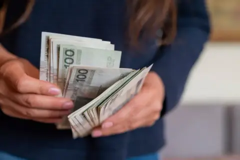 Dłonie trzymające plik banknotów 100 zł. Może to symbolizować wynagrodzenie lub zarobki w budżetówce, gdzie grupy zaszeregowania wpływają na pensję.