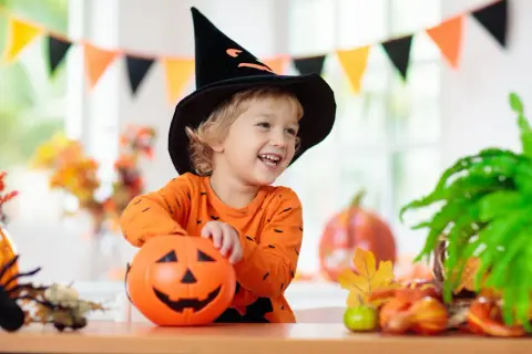 Uśmiechnięty chłopiec w czarnej, spiczastej czapce czarownicy i pomarańczowej bluzce z nietoperzami trzyma dynię. To idealny strój owoc dla dziecka na Halloween!