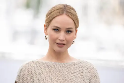 Jennifer Lawrence, gwiazda wielu filmów, w beżowym swetrze i złotych kolczykach.