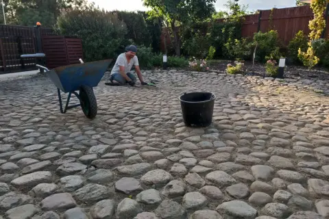 Mężczyzna pracuje przy podjeździe z kamienia polnego, obok stoi taczka i wiadro.