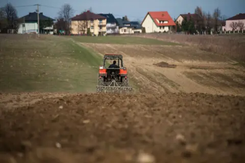 Traktor orze pole, przygotowując je pod przyszłą zabudowę. W tle widać domy, sugerujące warunki zabudowy na działce rolnej.