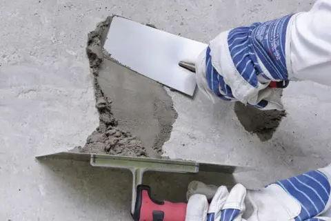 Naprawa betonu na zewnątrz: ręce w rękawicach nakładają zaprawę kielnią na uszkodzony fragment ściany.