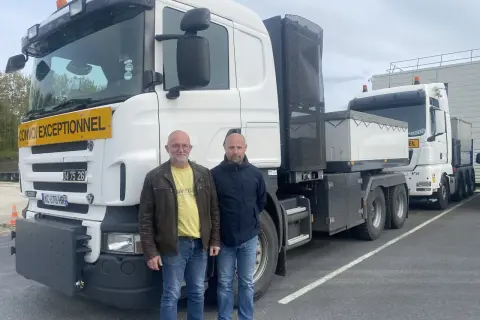 Deux chauffeurs poids lourd national posent devant leurs camions. L'un porte un gilet jaune, l'autre une veste bleue.