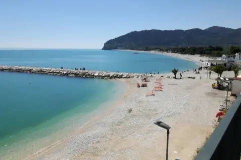 Mattinata serena sulla spiaggia di piana, con il mare azzurro che lambisce la costa ghiaiosa e il molo di sassi.