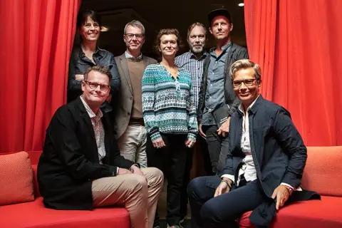 Gruppbild med åtta personer framför röda draperier. Izabella Jensen står i mitten, klädd i en mönstrad tröja.