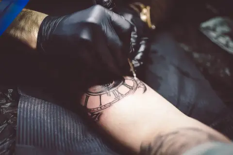 Artysta w czarnej rękawiczce tworzy tatuaż zegara z rzymskimi cyframi, stosując technikę whip shading dla uzyskania głębi.