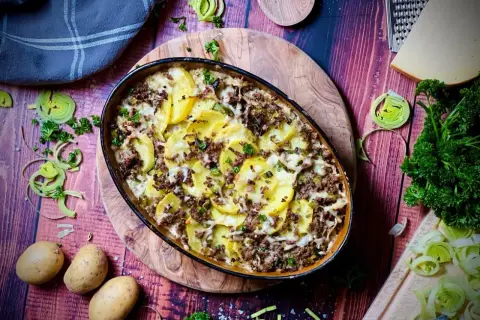 Herzhafter kartoffelauflauf mit hackfleisch, überbacken mit Käse und garniert mit Petersilie. Dazu frische Kartoffeln, Lauch und ein Stück Käse.