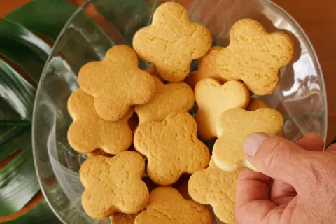 Una mano prende uno dei tanti biscotti fatti in casa a forma di quadrifoglio, disposti in una ciotola di vetro con foglie verdi sullo sfondo.