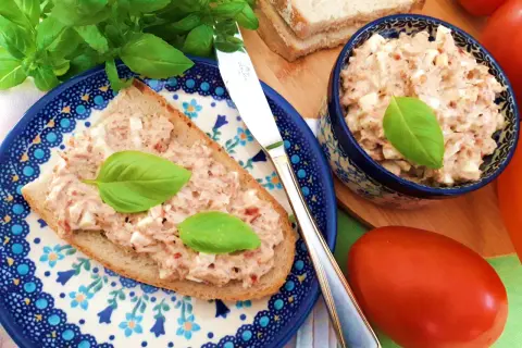 Pyszna pasta jajeczna z tuńczykiem na kromce chleba, ozdobiona bazylią. Obok miseczka z pastą i świeże pomidory.