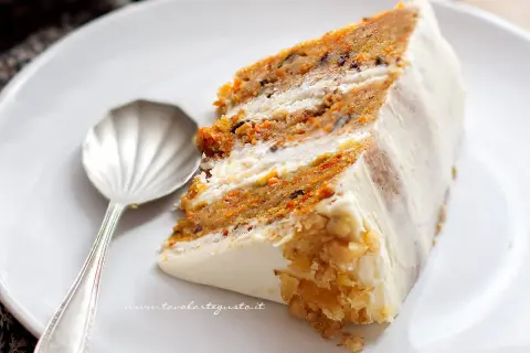 Una fetta di torta di carote, soffice e profumata, con glassa bianca e noci tritate. Accanto, un cucchiaino d'argento.