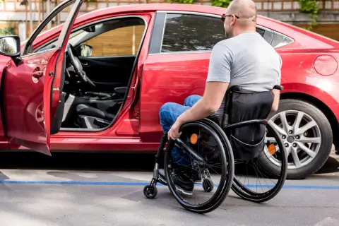 Uomo in carrozzina accanto a un'auto rossa, con portiera aperta. Un esempio di accessibilità, come previsto dall'art. 30 comma 7 legge 388 del 2000.