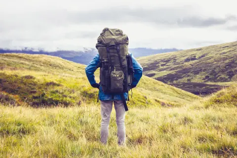 Wanderer mit großem Rucksack steht auf einem Hügel, bereit für seine 1 Jahr Weltreise. Die Landschaft ist grün und hügelig.