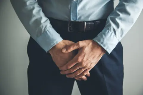 Un homme en chemise bleue et pantalon sombre, mains croisées devant lui, semble avoir un testicule qui gratte.