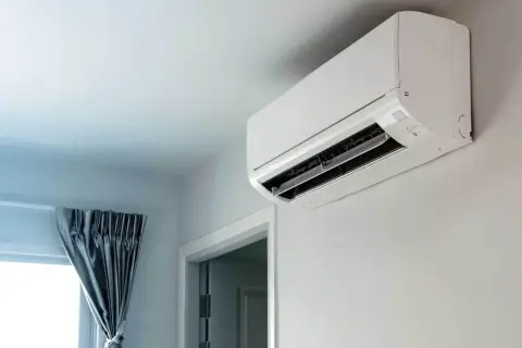 Biała jednostka klimatyzacyjna zamontowana na jasnej ścianie, obok drzwi i okna z zasłonami.