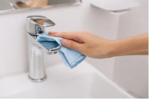 Mano limpiando el grifo cromado del lavabo con un paño azul. La limpieza del baño es esencial para un hogar saludable.