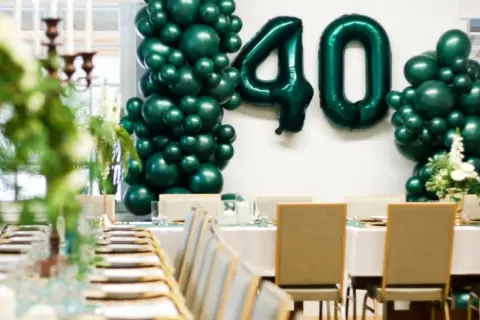 Uroczyste przyjęcie z okazji 40 urodzin. Zielone balony tworzą liczbę 40. Stół nakryty do eleganckiej kolacji.
