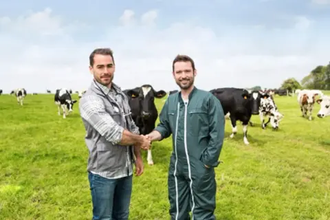 Due uomini si stringono la mano in un prato verde con tanti animali al pascolo.